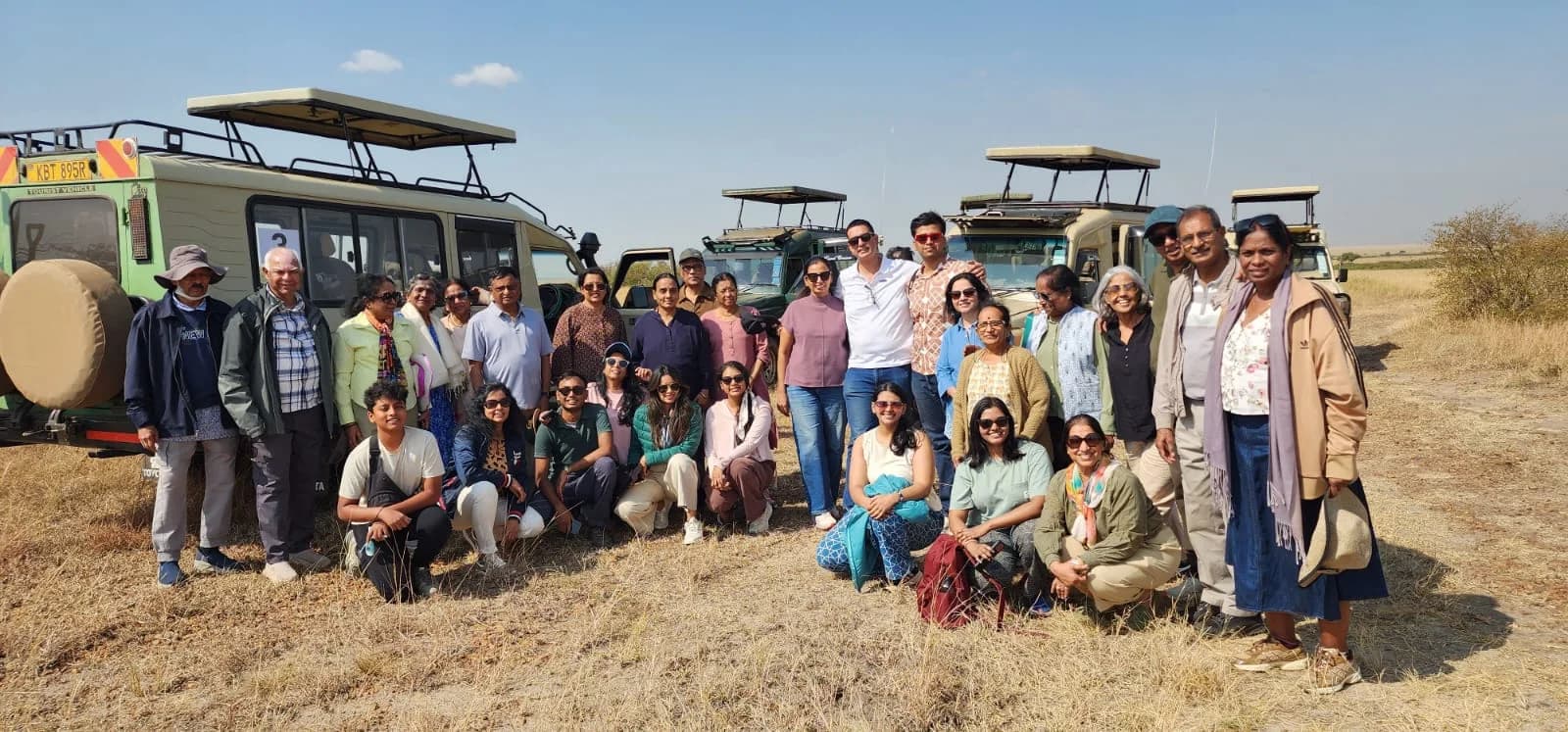 A large family group of all ages standing together beside safari vehicles on the plains